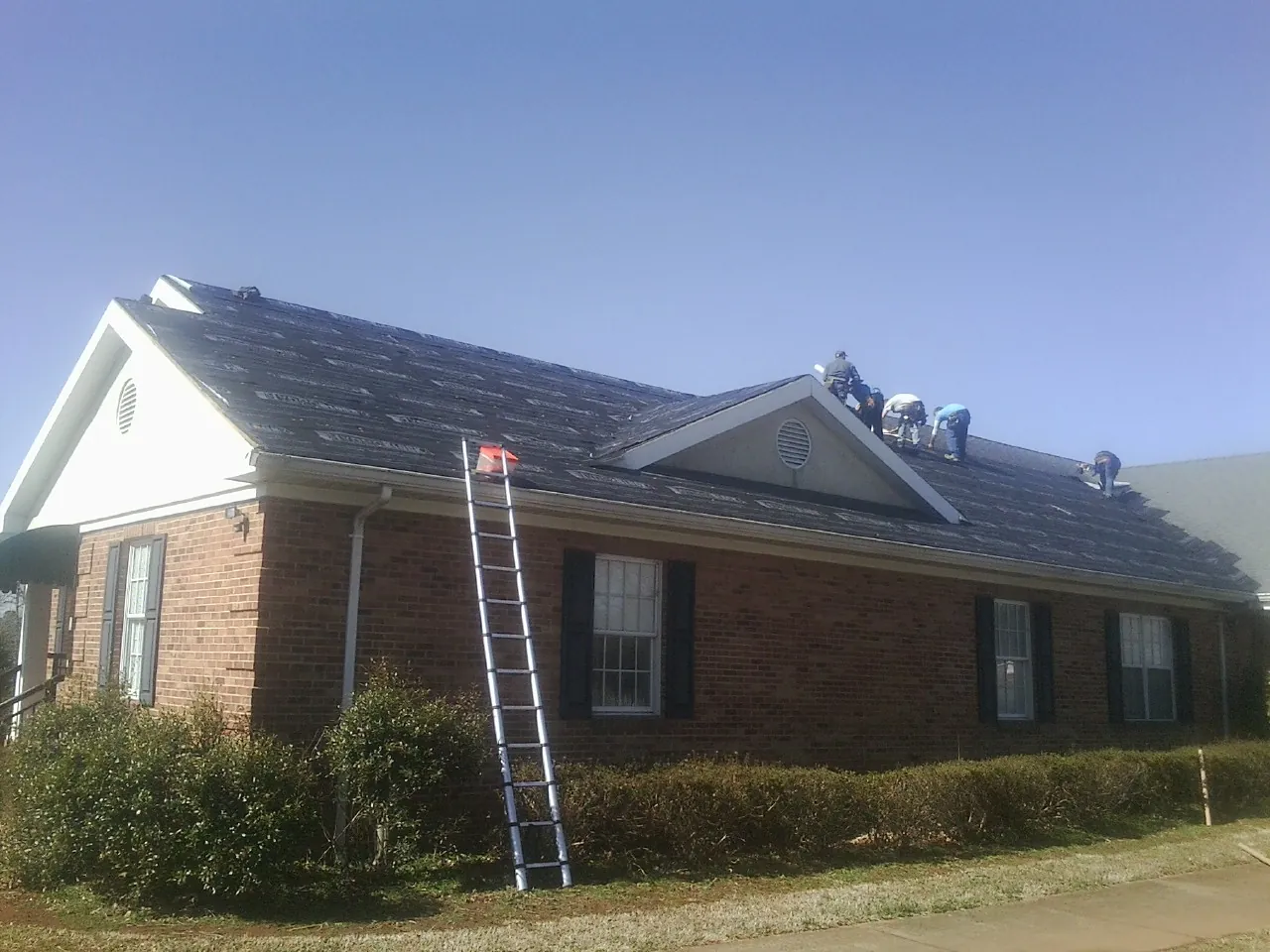 Crew tearing shingles off a brick ranch roof with a ladder set — Emergency Roof Repair work by our crew in Holt