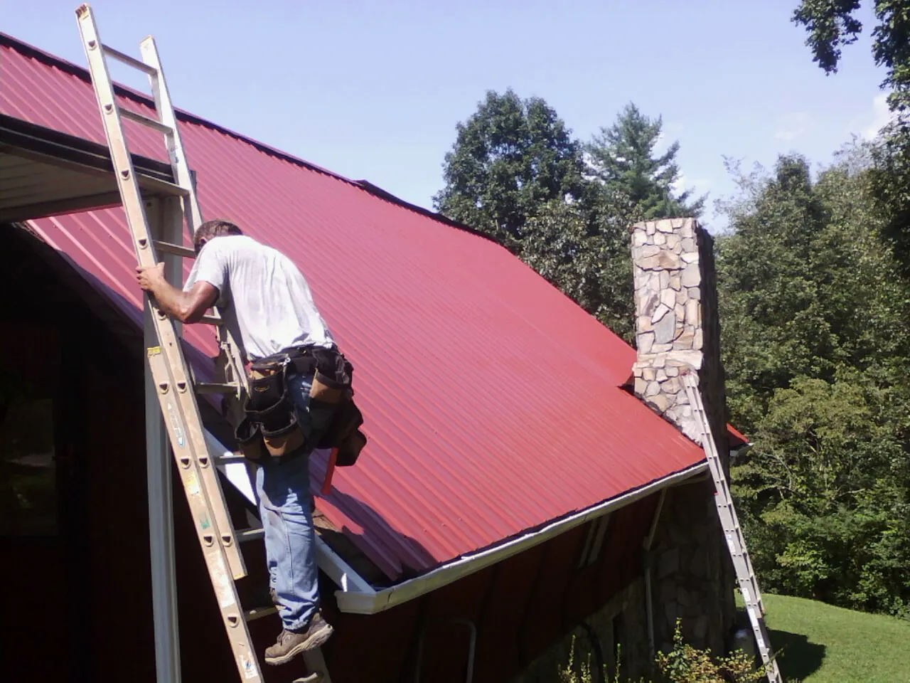 Roofer on a ladder beside a red metal roof and stone chimney — Soffit & Fascia Repair by Roofing Brothers in Holt